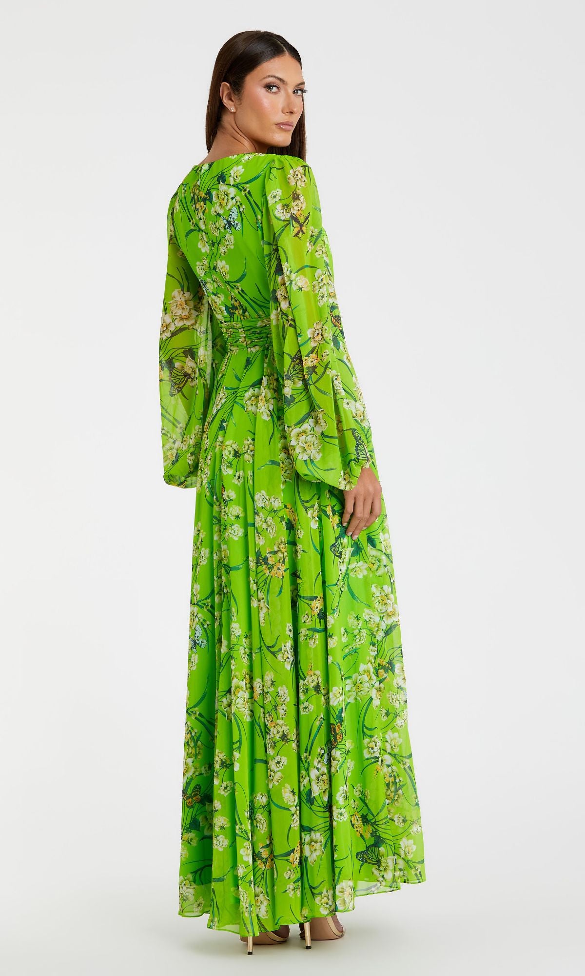 A woman in the Mac Duggal 56013 long green print formal dress with bright florals and long sleeves stands facing away, looking over her shoulder.